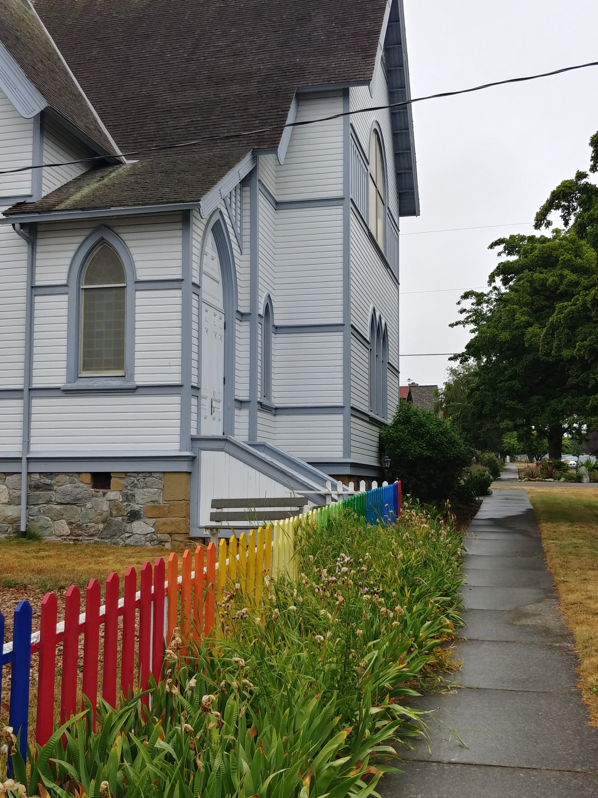 Side view of a light-blue wooden church with arched windows and stone foundation, next to a rainbow-colored picket fence and a sidewalk.