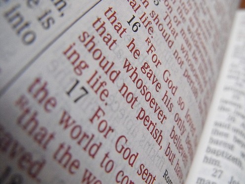 Close-up of a page with red text from a religious book; visible line reads 'For God so loved the world'.
